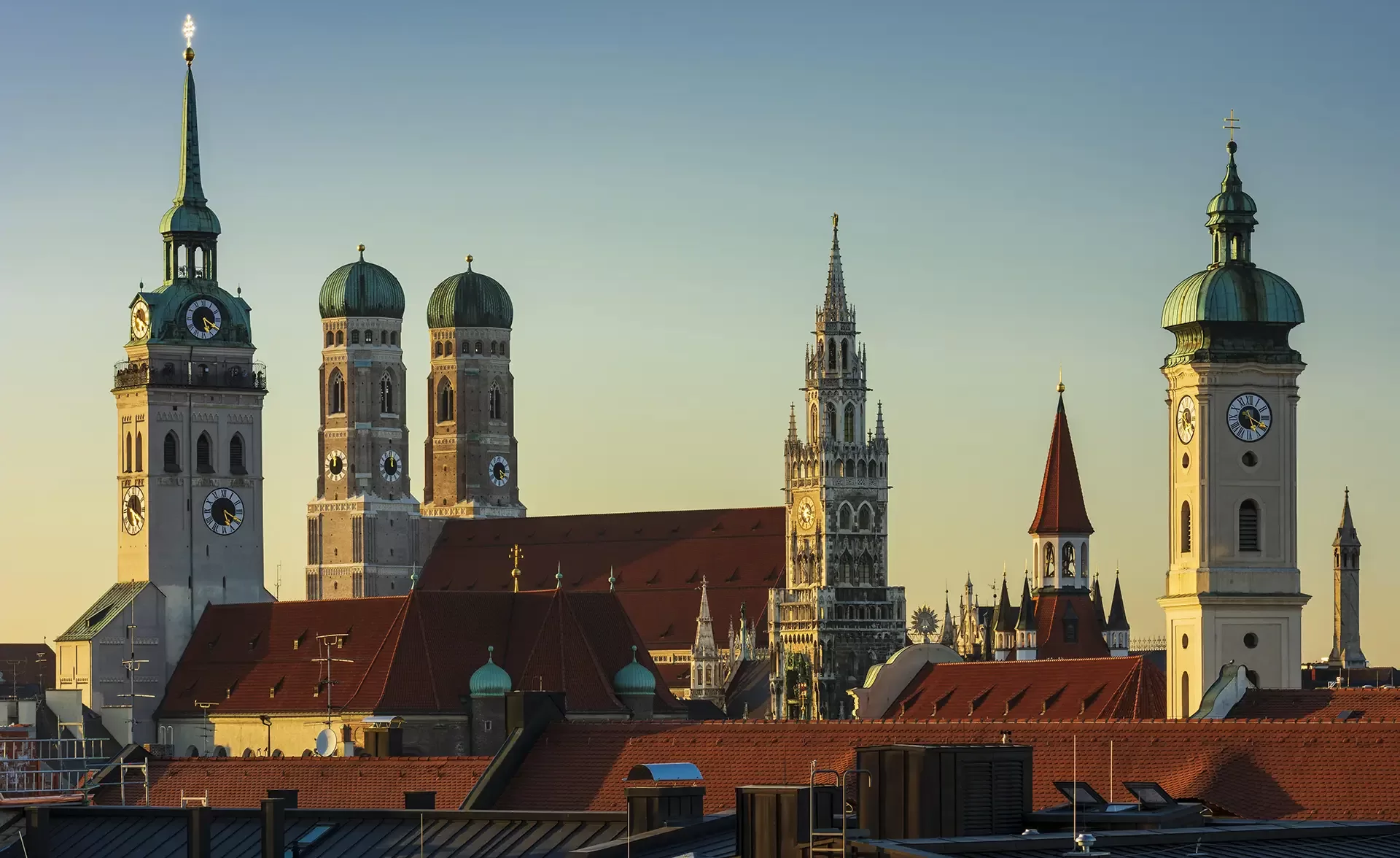 Munich cityscape at sunset, featuring the Frauenkirche twin domed towers, New Town Hall with its clock tower, and other historic buildings with red rooftops against a clear sky.