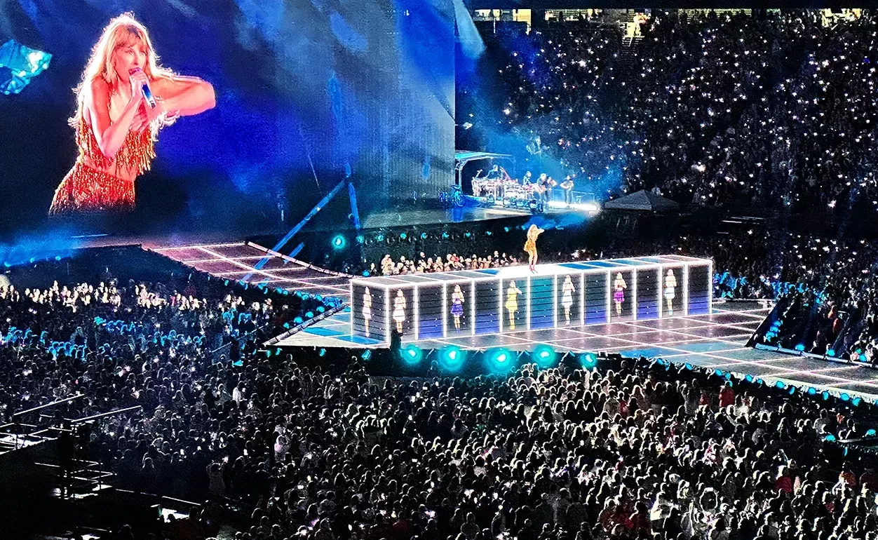 A singer performs on an illuminated stage set above a large, enthusiastic crowd in a stadium. A giant screen displays a close-up of the performer wearing a shimmering outfit. The audience is bathed in colorful lights.