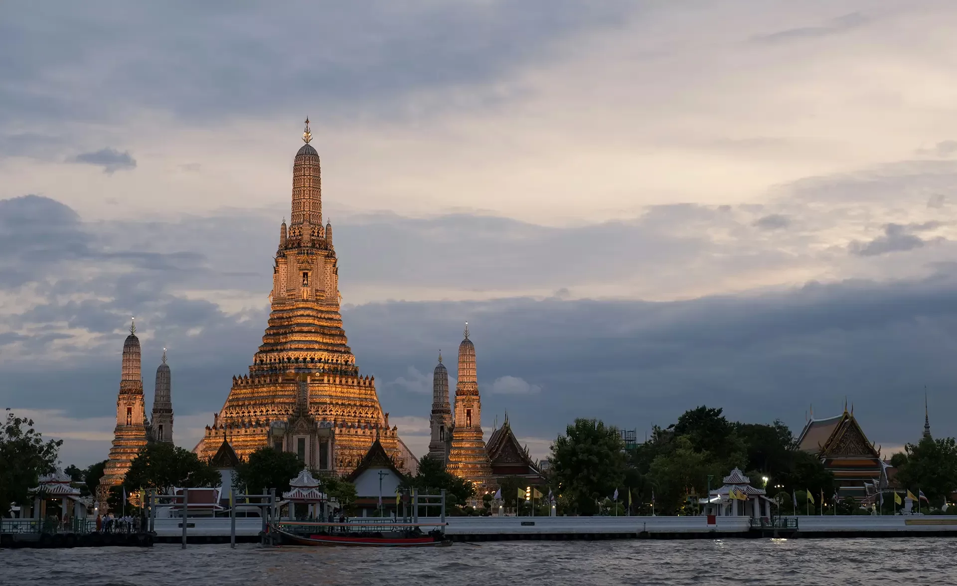 Wat Arun temple in Bangkok, Thailand, at dusk with the central spire illuminated. Clouds fill the sky, and a river with moored boats is in the foreground, adding to the serene atmosphere.