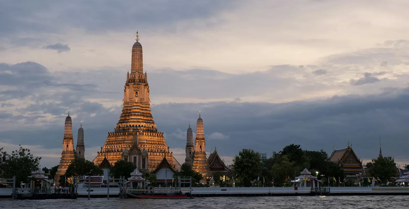 The image shows the Wat Arun temple in Bangkok, Thailand, illuminated at dusk. The temples intricate architecture is reflected in the calm river in the foreground, with a cloudy sky in the background.