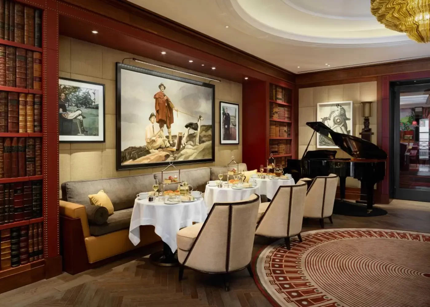 Elegant restaurant with round tables set for afternoon tea, plush chairs, a grand piano, bookshelves, and large framed art on the walls, creating a sophisticated and cozy ambiance.