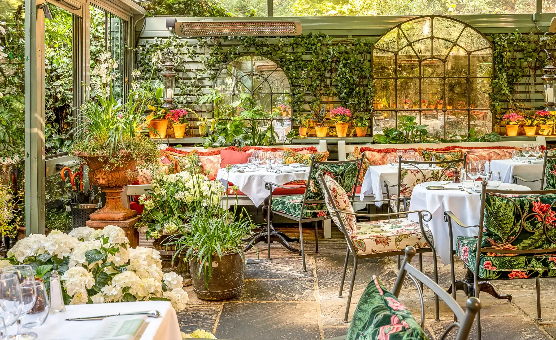 A lush, sunny conservatory restaurant with tables set for dining. The space features abundant plants, floral-patterned chairs, white tablecloths, and large windows. Greenery and flowers create a vibrant, inviting atmosphere.