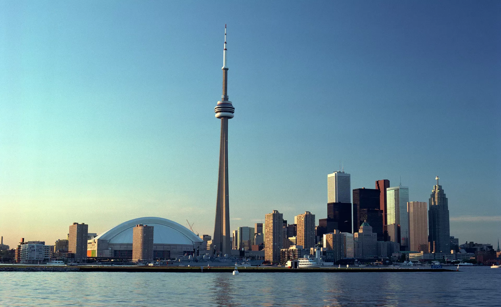 The image shows the skyline of Toronto with the CN Tower prominently in the center. Surrounding the tower are various skyscrapers, and the Rogers Centre with its domed roof. The scene is reflected in the calm water in the foreground.