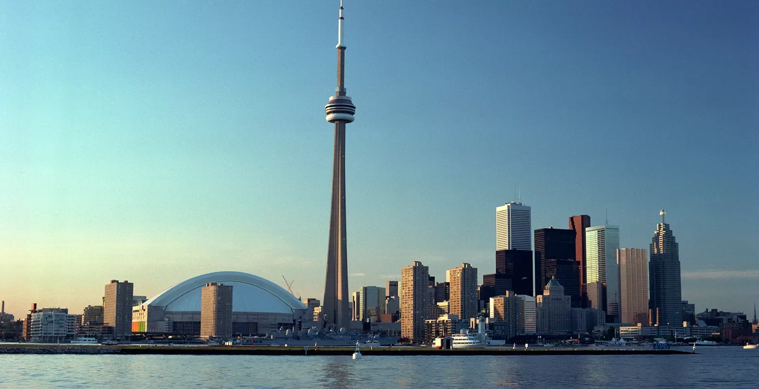 Toronto skyline featuring the CN Tower prominently against a clear blue sky. The Rogers Centre is visible to the left. The citys modern skyscrapers are reflected in the calm waters of Lake Ontario.