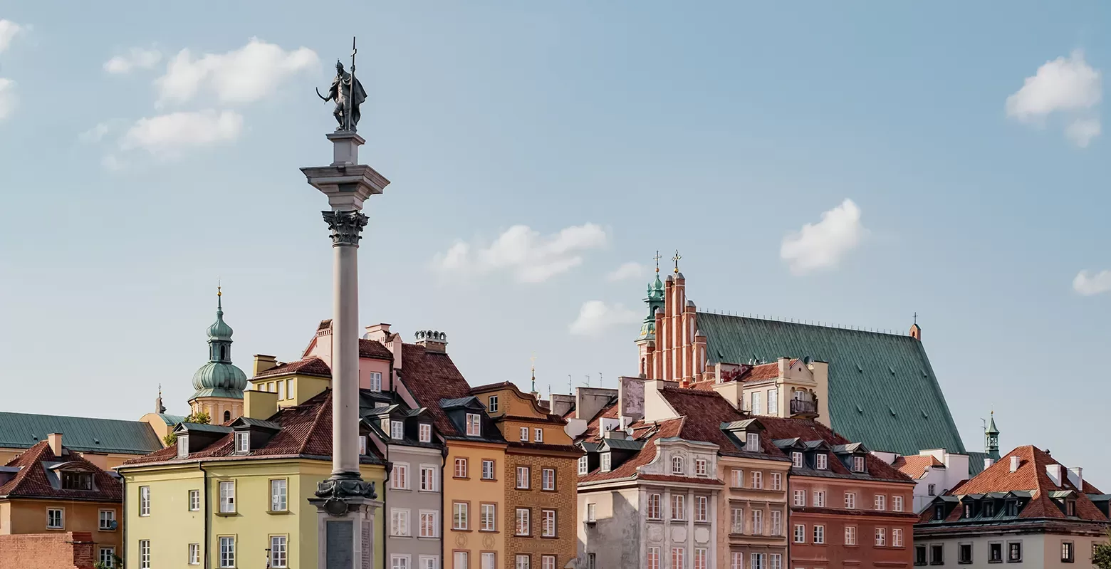 A scenic view of colorful historic buildings in a European city, featuring a prominent column topped with a statue. The sky is clear with a few clouds, and traditional architecture dominates the landscape, creating a picturesque urban scene.