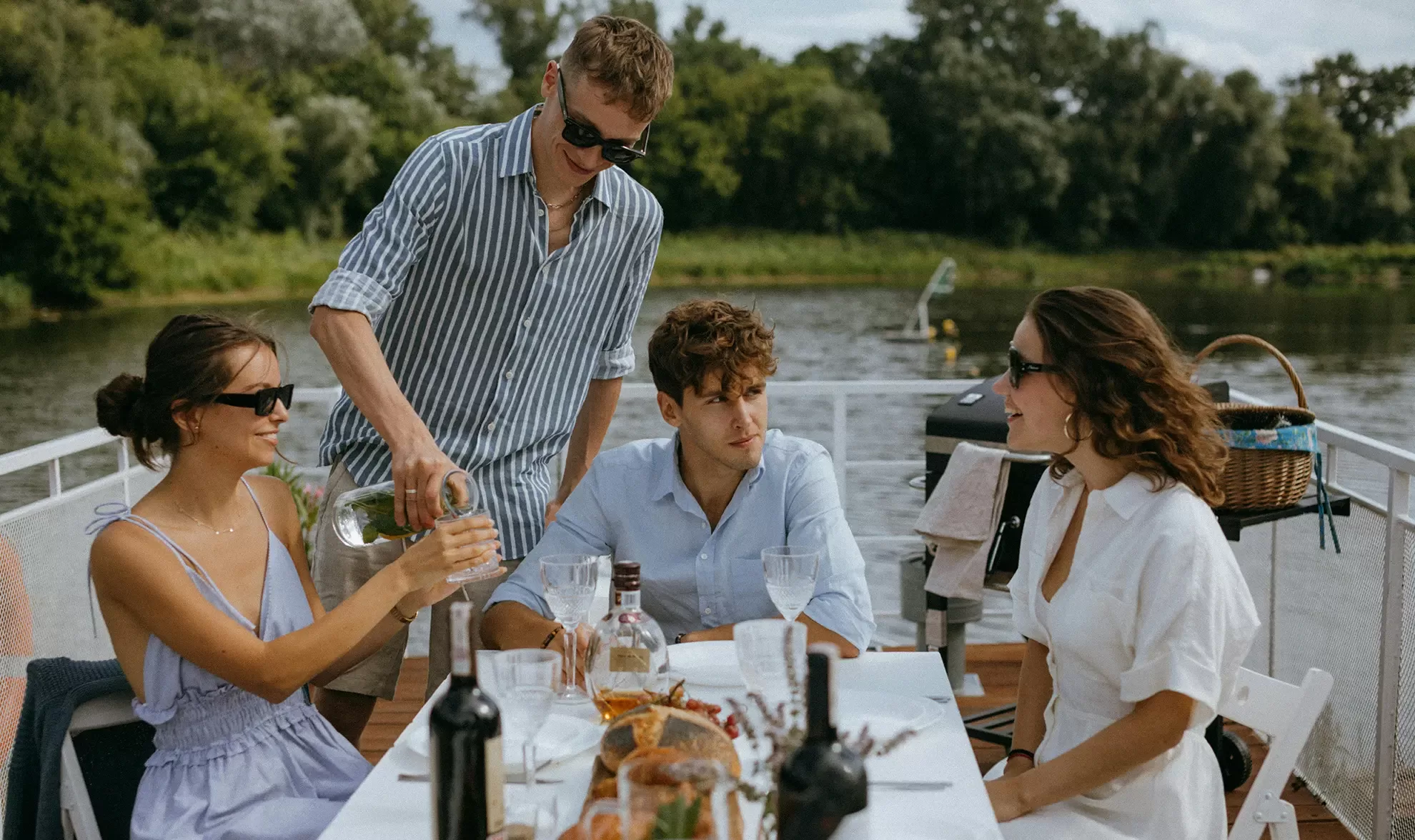 Four people sit around a table on a boat outdoors, dressed in light summer clothes and sunglasses. One person pours a drink while others smile and chat. Food, wine, and glassware are on the table. Trees and water are in the background.